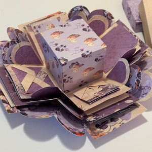 May include: A purple and brown explosion box with a floral pattern. The box is open and has multiple layers. The box is decorated with a small square box with a floral pattern.