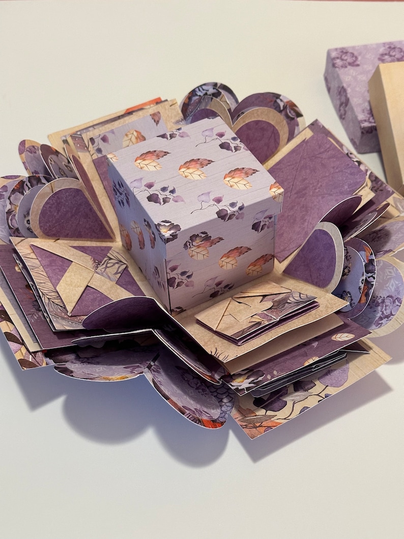 May include: A purple and brown explosion box with a floral pattern. The box is open and has multiple layers. The box is decorated with a small square box with a floral pattern.