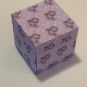May include: A small, square gift box with a floral pattern in shades of purple and white. The box is made of cardboard and has a hinged lid.
