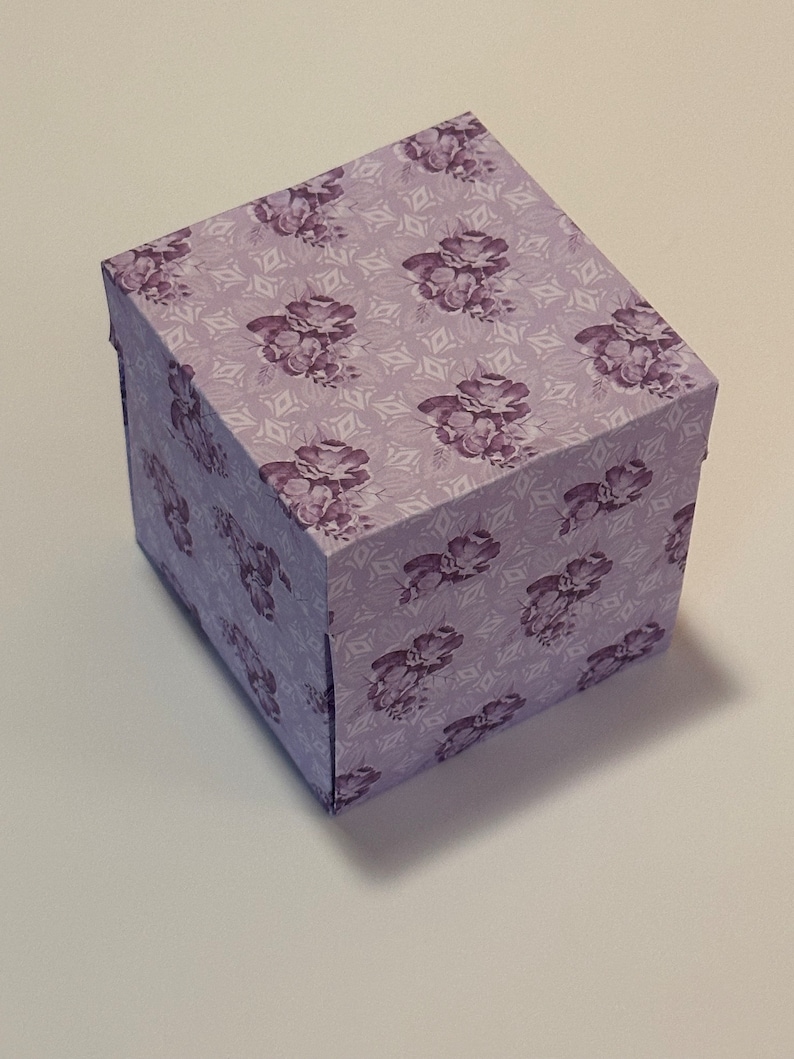 May include: A small, square gift box with a floral pattern in shades of purple and white. The box is made of cardboard and has a hinged lid.
