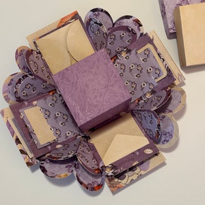 May include: A purple explosion box with a floral pattern and multiple layers. The box is open and has a small square box in the center. The box is decorated with a floral pattern and has a scalloped edge.