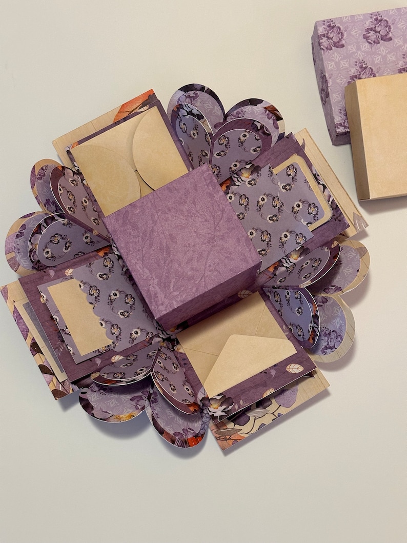 May include: A purple explosion box with a floral pattern and multiple layers. The box is open and has a small square box in the center. The box is decorated with a floral pattern and has a scalloped edge.