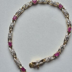 May include: A gold and silver-toned bracelet featuring oval-cut pink rubies alternating with X-shaped metal links. The links are accented with small, clear stones. The bracelet includes a secure box clasp closure with a safety latch mechanism.