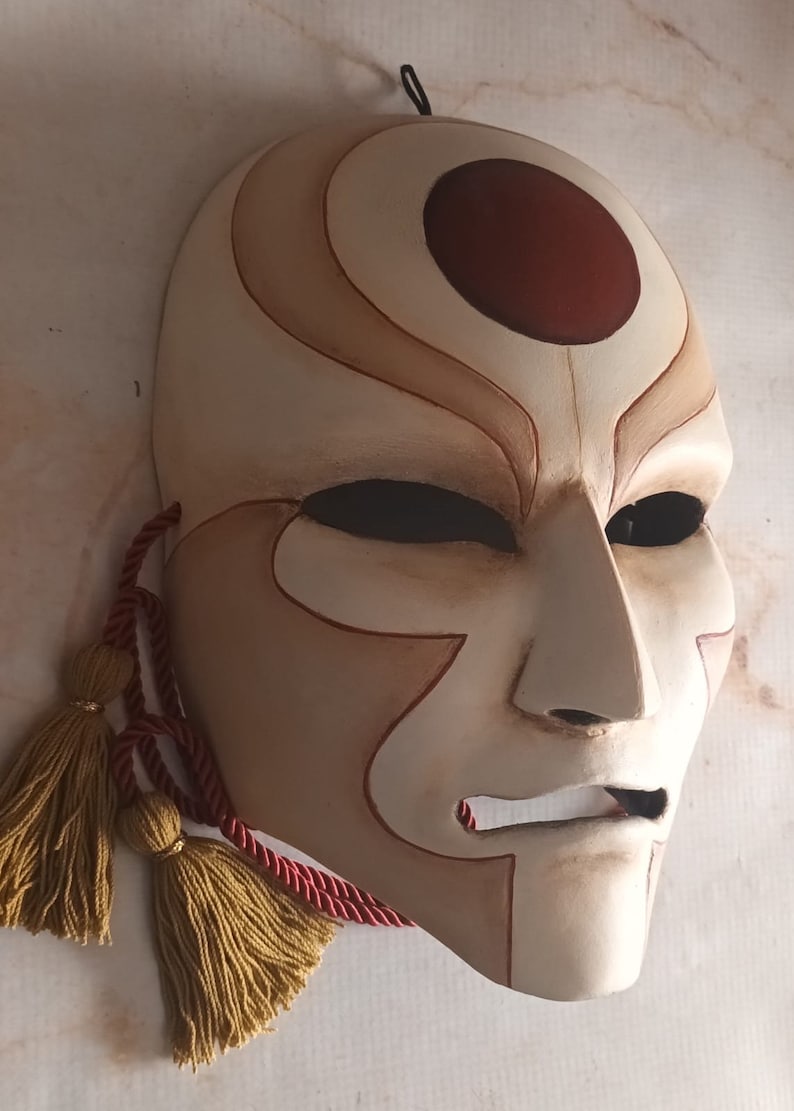 Pode incluir: Uma m&aacute;scara branca com um c&iacute;rculo vermelho e linhas marrons. A m&aacute;scara tem um rosto humano com a boca e os olhos fechados. A m&aacute;scara est&aacute; presa a um cord&atilde;o vermelho com borlas douradas.