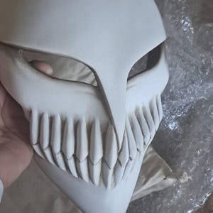 May include: A white, stylized mask with a prominent jawline and sharp teeth. The mask has a smooth, matte finish and is held by a hand. The design features a large, open mouth and eye openings, suggesting a character from a fantasy or anime series.