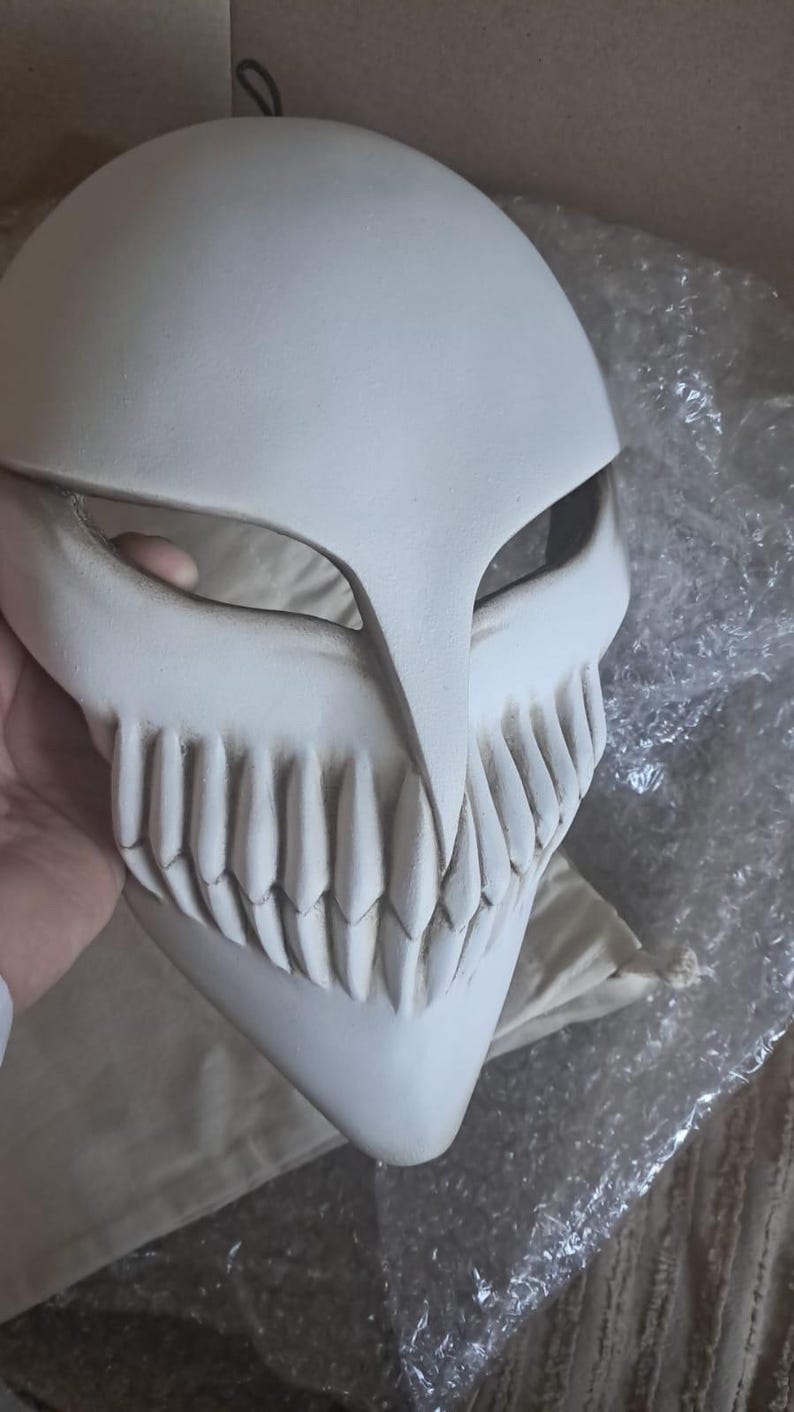 May include: A white, stylized mask with a prominent jawline and sharp teeth. The mask has a smooth, matte finish and is held by a hand. The design features a large, open mouth and eye openings, suggesting a character from a fantasy or anime series.