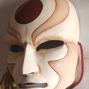 Pode incluir: Uma m&aacute;scara branca com detalhes pintados em vermelho e marrom. A m&aacute;scara tem um c&iacute;rculo vermelho grande na testa e um rosto estilizado com uma boca pequena e dois buracos para os olhos. A m&aacute;scara est&aacute; presa a um cord&atilde;o vermelho.