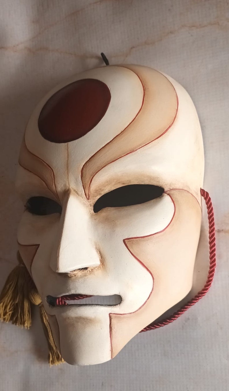 Pode incluir: Uma m&aacute;scara branca com detalhes pintados em vermelho e marrom. A m&aacute;scara tem um c&iacute;rculo vermelho grande na testa e um rosto estilizado com uma boca pequena e dois buracos para os olhos. A m&aacute;scara est&aacute; presa a um cord&atilde;o vermelho.