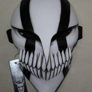 May include: A white and black striped mask with a menacing design. The mask features sharp teeth and a pointed chin. A black strap is attached to the sides. A tag with Japanese text hangs from the mask.
