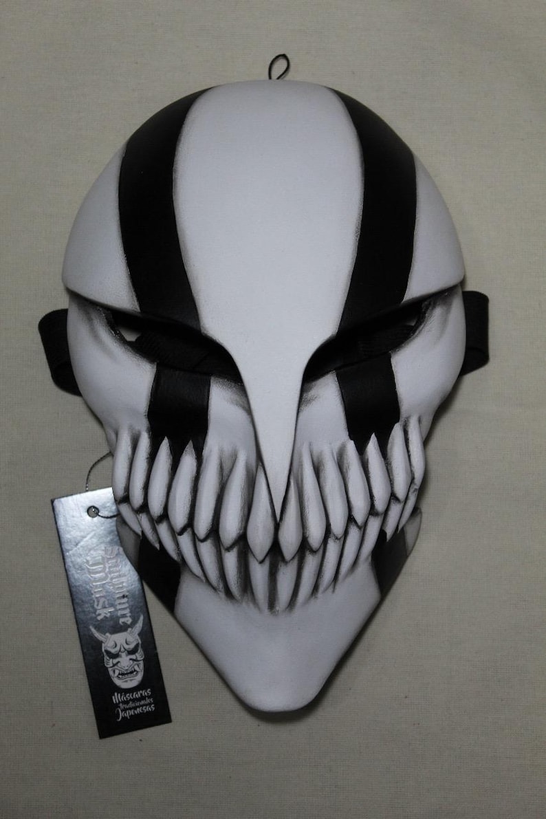 May include: A white and black striped mask with a menacing design. The mask features sharp teeth and a pointed chin. A black strap is attached to the sides. A tag with Japanese text hangs from the mask.
