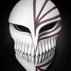 May include: A white mask with a skull-like design, featuring sharp teeth and dark red markings. The mask has a pointed chin and eye openings, with red lines radiating from the forehead. The overall aesthetic is reminiscent of a fantasy or anime theme.