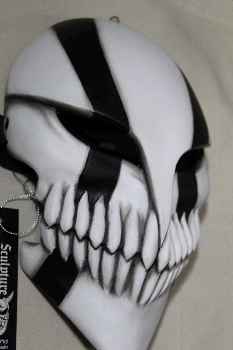 May include: A white and black mask with a skull-like design. The mask features black stripes and sharp, pointed teeth. The mask has a tag that says "Sculpture".