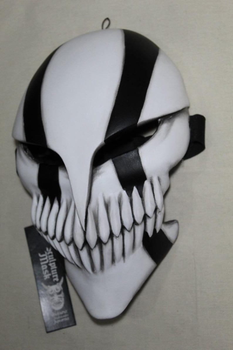 May include: A white and black mask with a skull-like design. The mask features black stripes, sharp teeth, and a pointed beak-like structure. A black strap is attached to the side. A tag with the text "Sculpture Mask" is visible.