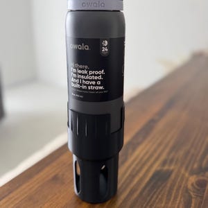 May include: A gray reusable water bottle with a black lid and a built-in straw. The bottle has a purple band with the word "owala" printed on it. The bottle is sitting on a wooden surface. The text on the bottle reads: "Hi there. I'm leak proof. I'm insulated. And I have a built-in straw."