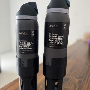 May include: Two gray reusable water bottles with black lids and built-in straws. The bottles have the text "owala" and "Hi there. I'm leak proof. I'm insulated. And I have a built-in straw." printed on them.