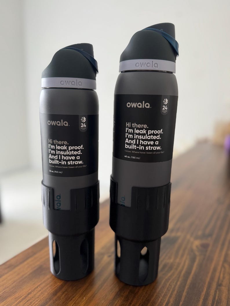 May include: Two gray reusable water bottles with black lids and built-in straws. The bottles have the text "owala" and "Hi there. I'm leak proof. I'm insulated. And I have a built-in straw." printed on them.