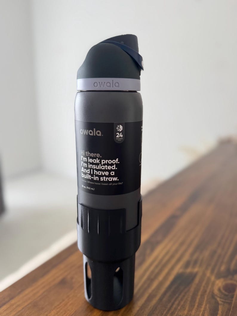May include: A gray reusable water bottle with a black lid and a built-in straw. The bottle has a purple band with the word "owala" printed on it. The bottle is sitting on a wooden surface. The text on the bottle reads: "Hi there. I'm leak proof. I'm insulated. And I have a built-in straw."