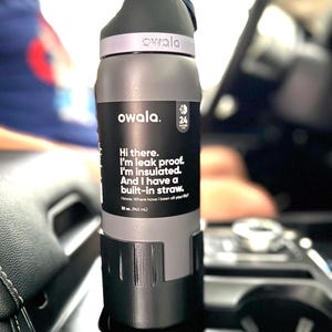 May include: A gray insulated water bottle with a built-in straw. The bottle has a black label with the text "owala." and "Hi there. I'm leak proof. I'm insulated. And I have a built-in straw. 24 hours cold. 12 oz. (345 mL)"