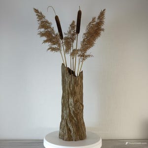 May include: A brown, tree trunk-shaped vase with a textured surface, holding dried cattails and ornamental grasses. The vase sits on a white pedestal. The cattails are dark brown. The vase is approximately 20 cm tall.