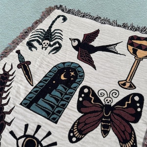 Puede incluir: Un tapiz tejido con una variedad de diseños de tatuajes tradicionales, que incluyen un escorpión, un pájaro, una mariposa, un ojo, una daga, un cáliz y una puerta. El tapiz es de color crema con acentos negros, marrones, rojos, azules y dorados.