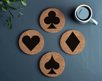 May include: Four round cork coasters with black card suit symbols: hearts, clubs, diamonds, and spades. The coasters are arranged on a dark blue surface, with a cup of coffee and greenery in the background. The coasters are a natural brown color.
