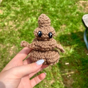 Op de afbeelding: Een bruin gehaakt poep-emoji pluche speelgoed met zwarte ogen. Het speelgoed wordt vastgehouden in een hand met witte nagellak.