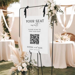 May include: A white wedding sign on a black metal stand with the text "YOUR SEAT awaits" and a QR code. The sign also includes the names "SOPHIA and ALEX" and the date "JULY 18, 2026". The sign is decorated with white flowers and greenery.