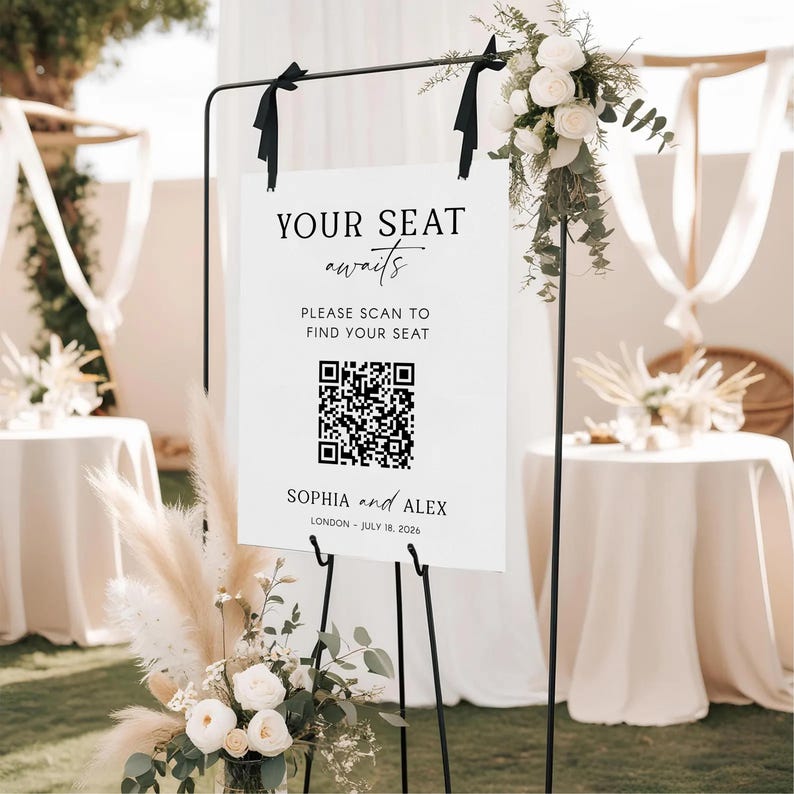 May include: A white wedding sign on a black metal stand with the text "YOUR SEAT awaits" and a QR code. The sign also includes the names "SOPHIA and ALEX" and the date "JULY 18, 2026". The sign is decorated with white flowers and greenery.