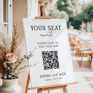 May include: A white sign on a wooden easel with the text "YOUR SEAT awaits" and "PLEASE SCAN TO FIND YOUR SEAT". Below is a QR code and the names "SOPHIA and ALEX" with the date "LONDON - JULY 18, 2026".