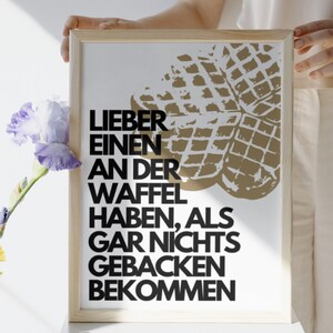 May include: A framed print with a white background and black text that reads "Lieber einen an der Waffel haben, als gar nichts gebacken bekommen". The print features a brown waffle graphic in the upper right corner.