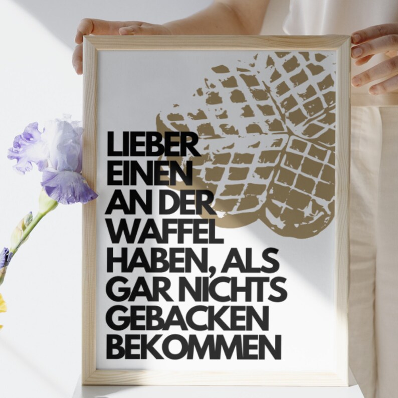 May include: A framed print with a white background and black text that reads "Lieber einen an der Waffel haben, als gar nichts gebacken bekommen". The print features a brown waffle graphic in the upper right corner.