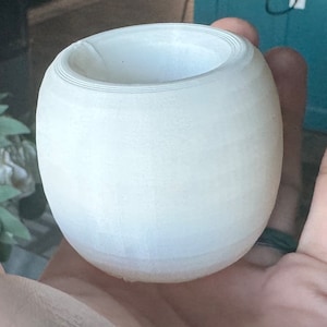 May include: A white, round, ceramic planter with a smooth, slightly textured surface. The planter has a wide opening at the top and a slightly rounded bottom.