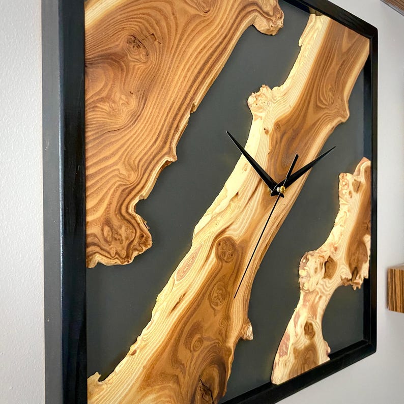 May include: Square wooden wall clock with a black frame. The clock face features natural wood grain patterns and a dark gray resin infill. Black clock hands indicate the time.