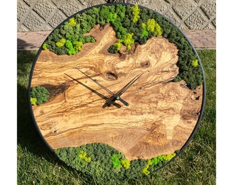 Könnte beinhalten: Eine runde Holzuhr mit schwarzem Metallrahmen. Das Zifferblatt der Uhr ist aus einer natürlichen Holzscheibe mit grünen Moosakzenten gefertigt. Die Uhrzeiger sind schwarz.
