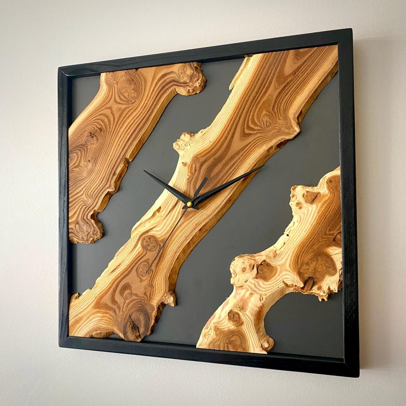 May include: Square wooden wall clock with a black frame. The clock face features natural wood grain patterns set against a dark gray background. Black clock hands indicate the time.