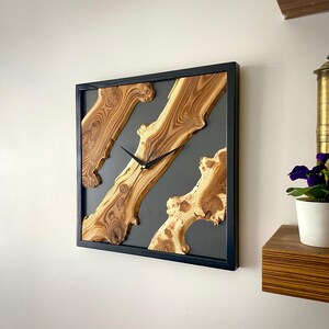 May include: Square wooden wall clock with a black frame. The clock face features natural wood grain patterns set against a gray background. Black clock hands indicate the time. A decorative piece for home decor.