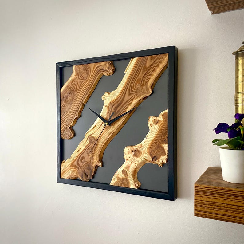 May include: Square wooden wall clock with a black frame. The clock face features natural wood grain patterns set against a gray background. Black clock hands indicate the time. A decorative piece for home decor.