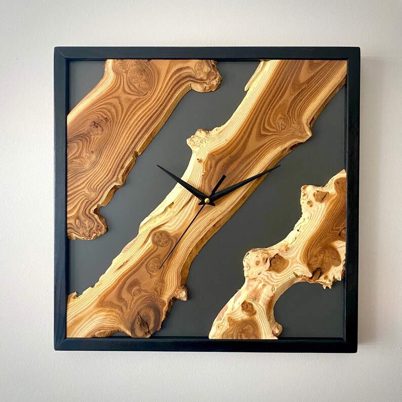 May include: Square wooden wall clock with a black frame. The clock face features natural wood grain patterns set against a dark gray background. Black clock hands indicate the time.
