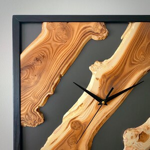 May include: Square wooden wall clock with a black frame. The clock face features natural wood grain patterns and black clock hands. The background is a dark gray color.