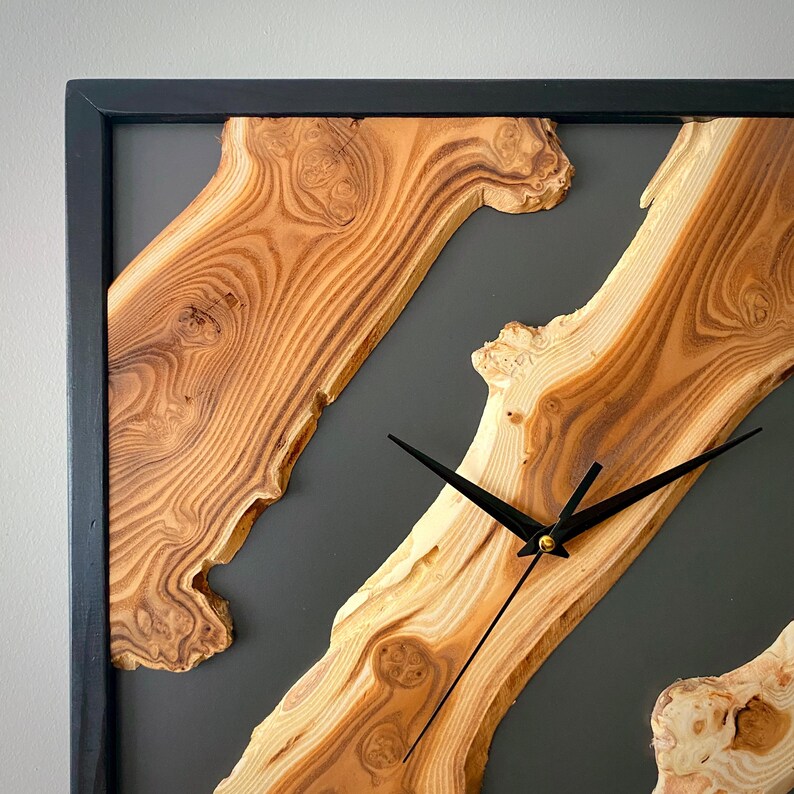 May include: Square wooden wall clock with a black frame. The clock face features natural wood grain patterns and black clock hands. The background is a dark gray color.