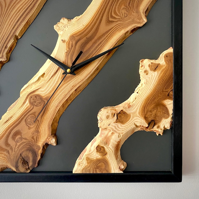 May include: Square wooden wall clock with a black frame. The clock face features natural wood grain patterns and black clock hands. The background is a dark gray color.