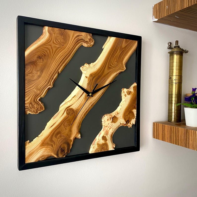 May include: Square wooden wall clock with a black frame. The clock face features natural wood grain patterns set against a dark gray background. Black clock hands indicate the time. A unique, decorative home decor item.