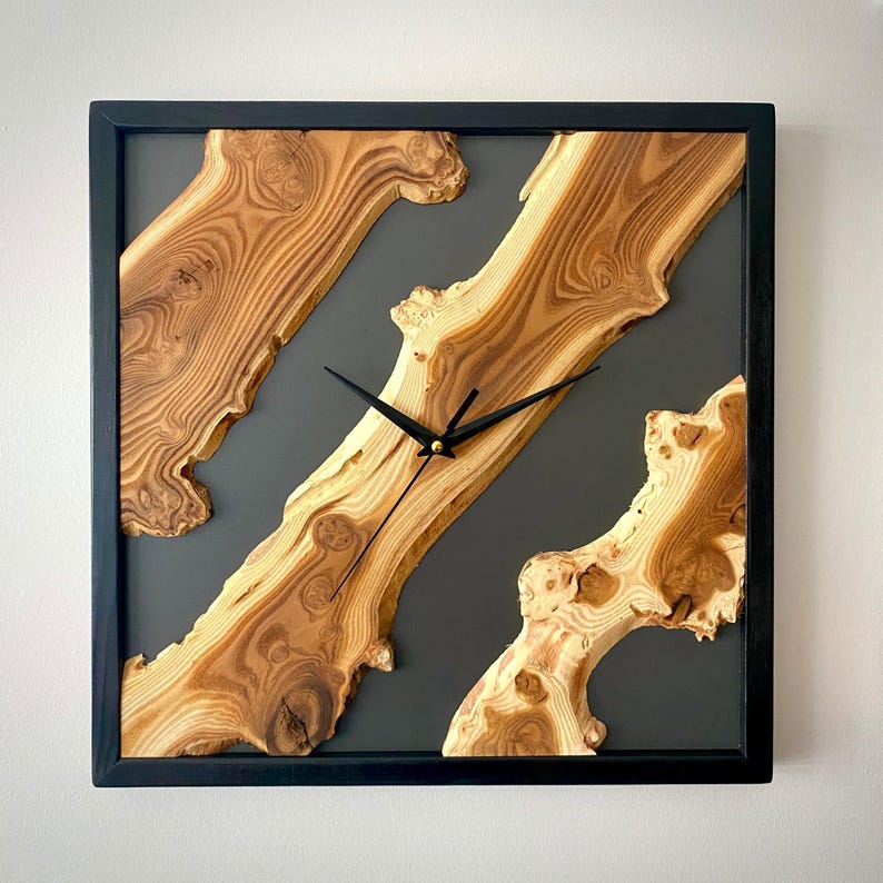 May include: Square wooden wall clock with a black frame. The clock face features natural wood grain patterns in shades of brown and tan, set against a dark gray background. Black clock hands indicate the time.