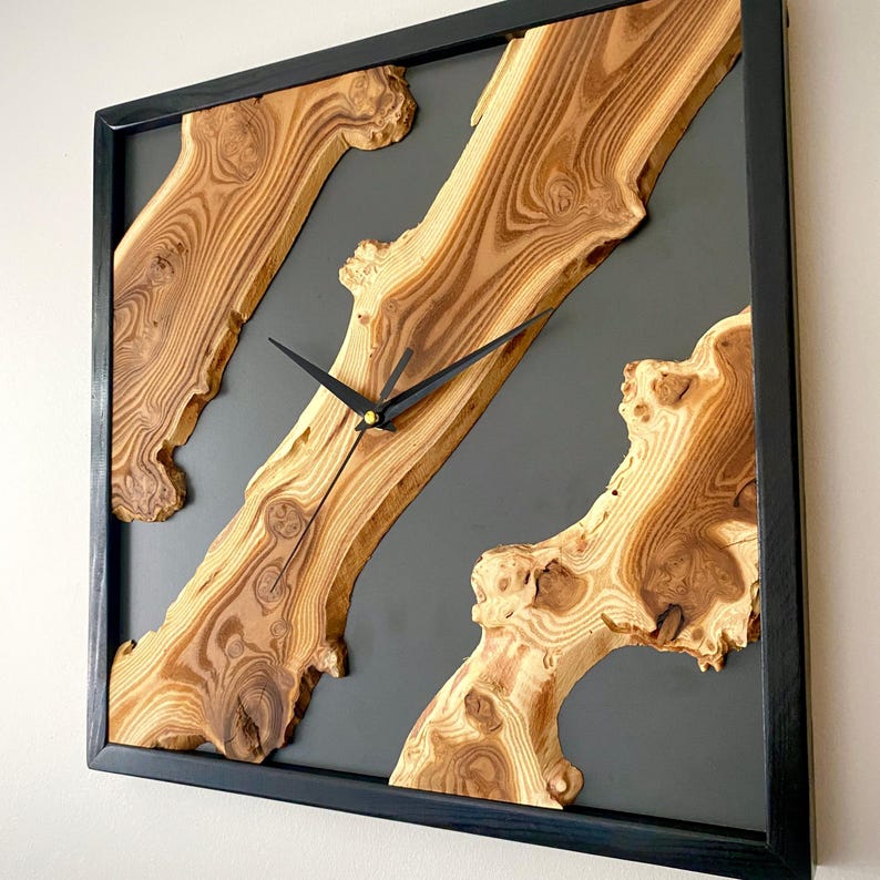 May include: Square wooden wall clock with a black frame. The clock face features natural wood grain patterns set against a gray background. Black clock hands indicate the time. A unique, rustic home decor item.