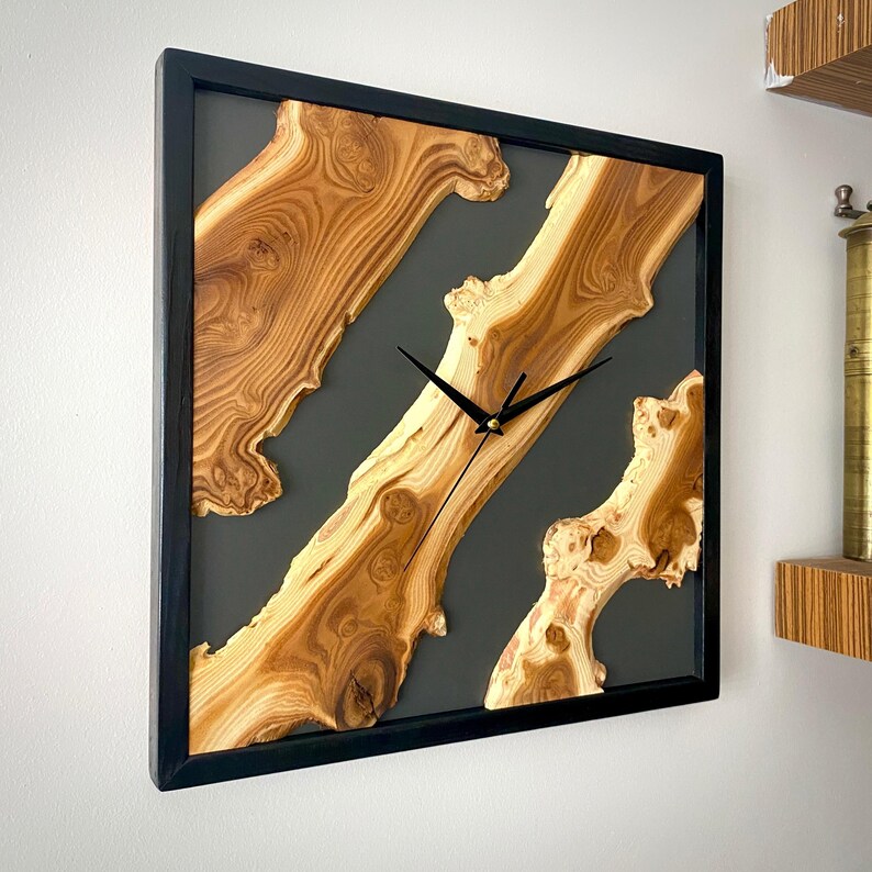 May include: Square wooden wall clock with a black frame. The clock face features natural wood grain patterns in shades of brown and tan, set against a dark gray background. Black clock hands indicate the time.