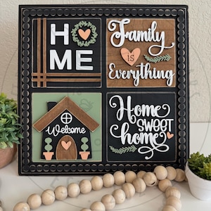 May include: A decorative wooden sign with a black frame and four square panels. The panels feature the words "HOME", "Family is Everything", "Welcome", and "Home Sweet Home" in white and brown lettering, with heart and leaf accents. The sign is a home decor item.