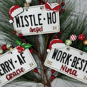 May include: Three white Christmas ornaments shaped like license plates with red and black text. The ornaments feature the phrases "Mistle Ho", "Merry-AF", and "Work-Besti" with names "Angel", "Grace", and "Nina" written below. Each ornament has a red Santa hat and a gold jingle bell.