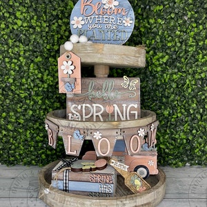 May include: A three-tiered wooden tray decorated with spring-themed decorations. The top tier features a round sign that says "Bloom Where You Are Planted". The middle tier has a sign that says "Hello Spring" and a banner that says "Bloom". The bottom tier has a sign that says "Spring Has Sprung" and a small truck with flowers.