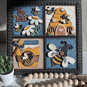 May include: A decorative wooden wall art piece featuring a bee theme. The artwork is divided into four squares, each with a different bee-related design, including a beehive, honey pot, and bees. The phrase "Bee Kind" is written in cursive.