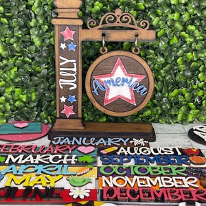 May include: Wooden sign with a red, white, and blue star and the word "America" on it. The sign is hanging from a wooden post with the word "July" and stars on it. There are also wooden signs with the months of the year on them.